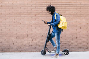 Junger Mann mit Rucksack und E-Scooter steht vor einer Backsteinmauer und schaut auf sein Handy - JRFF03366