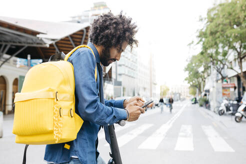 Mann mit Rucksack und E-Scooter bei der Nutzung einer Navigations-App auf seinem Mobiltelefon in der Stadt, Barcelona, Spanien - JRFF03371