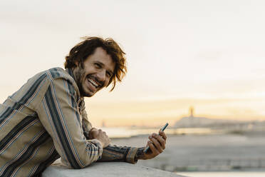 Portrait of laughing man with smartphone during sunset, Barcelona, Spain - AFVF03327