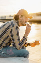 Tätowierter Mann mit Kaffee zum Mitnehmen, der bei Sonnenuntergang einen Krapfen isst - AFVF03341