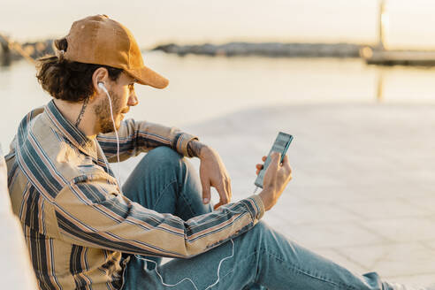 An der Uferpromenade sitzen und mit Kopfhörern und Smartphone bei Sonnenuntergang Musik hören - AFVF03345
