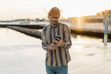Mann mit Baseballmütze hört Musik mit Kopfhörern und Smartphone bei Sonnenuntergang - AFVF03347