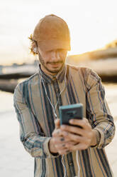 Man wearing baseball cap listening music with earphones and smartphone at sunset taking selfie - AFVF03349