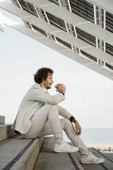 Geschäftsmann mit Kopfhörern sitzt auf einer Treppe im Freien und trinkt einen Kaffee zum Mitnehmen, Barcelona, Spanien - AFVF03365