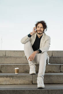 Porträt eines Geschäftsmannes mit Kaffee zum Mitnehmen, der auf einem Staris sitzt und mit Kopfhörern Musik hört - AFVF03366