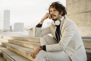 Portrait of businessman with headphones sitting on stairs outdoors looking at distance, Barcelona, Spain - AFVF03368