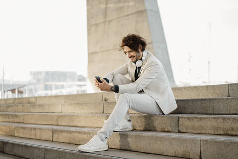 Lächelnder Geschäftsmann mit Kopfhörern, der auf einer Treppe im Freien sitzt und auf sein Mobiltelefon schaut, lizenzfreies Stockfoto