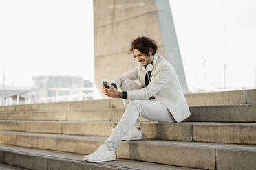 Lächelnder Geschäftsmann mit Kopfhörern, der auf einer Treppe im Freien sitzt und auf sein Mobiltelefon schaut - AFVF03369