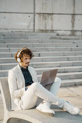 Businessman with headphones working on laptop outdoors - AFVF03378