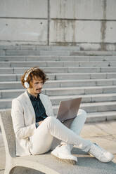 Geschäftsmann mit Kopfhörern bei der Arbeit am Laptop im Freien - AFVF03378