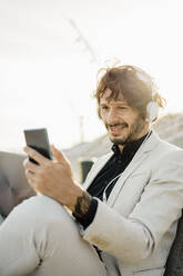 Portrait of smiling businessman with headphones and laptop looking at his mobile phone - AFVF03381