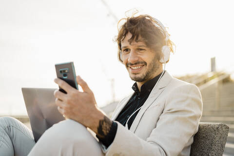 Porträt eines lächelnden Geschäftsmannes mit Kopfhörern und Laptop, der auf sein Mobiltelefon schaut, lizenzfreies Stockfoto