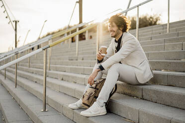 Laughing businessman with coffee to go and backpack sitting on stairs outdoors - AFVF03400