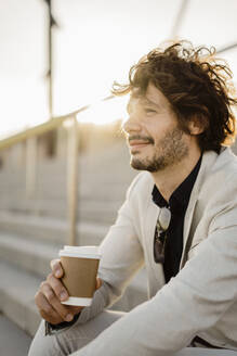 Lächelnder Geschäftsmann mit Kaffee zum Mitnehmen, der auf einer Treppe im Freien sitzt und sich entspannt - AFVF03401