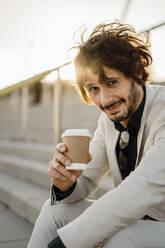 Porträt eines Geschäftsmannes mit Kaffee zum Mitnehmen auf einer Treppe sitzend bei Gegenlicht - AFVF03402