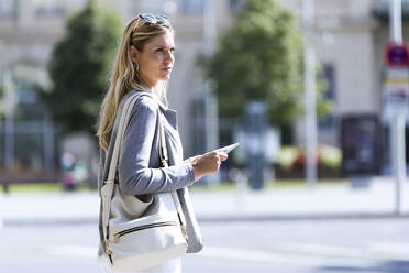 Junge Geschäftsfrau, die mit einem digitalen Tablet arbeitet, während sie mit einem Koffer auf der Straße steht - JSRF00281