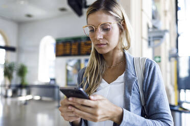 Junge Frau, die in der Bahnhofshalle mit ihrem Mobiltelefon SMS schreibt - JSRF00299