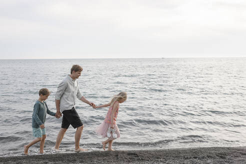 Vater geht Hand in Hand mit kleiner Tochter und Sohn am Meer spazieren - EYAF00255