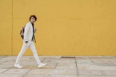 Portrait of walking businessman with backpack in front of yellow wall - AFVF03409