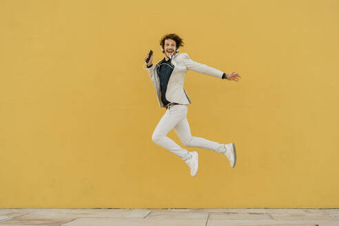 Happy businessman jumping in the air in front of yellow wall listening music with headphones - AFVF03416