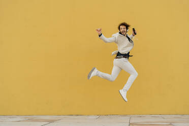 Businessman jumping in the air in front of yellow wall listening music with headphones and smartphone - AFVF03417