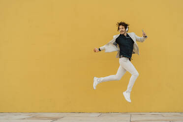 Businessman jumping in the air in front of yellow wall listening music with headphones and smartphone - AFVF03418
