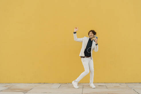Geschäftsmann singt und tanzt vor einer gelben Wand und hört Musik mit Kopfhörern und Smartphone - AFVF03420