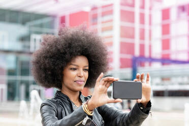 Junge Frau mit Afrofrisur an einer S-Bahn-Station, die ein Smartphone-Selfie macht - CUF51463