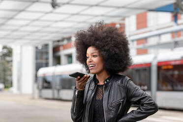 Junge Frau mit Afrofrisur an einer S-Bahn-Station, die mit einem Smartphone spricht - CUF51465