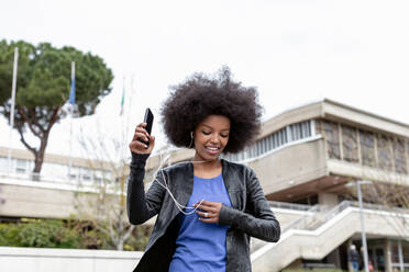 Junge Frau mit Afro-Haar in der Stadt, die spazieren geht und Smartphone-Musik hört - CUF51467