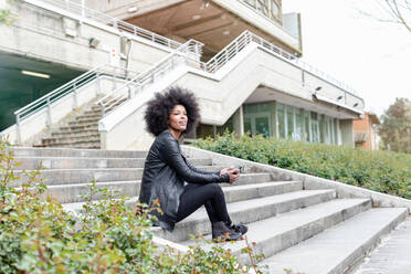 Junge Frau mit Afro-Haar sitzt auf einer Stadttreppe und schaut weg - CUF51469