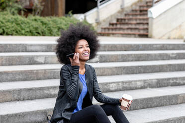 Happy young woman with afro hair sitting on city stairway, listening to smartphone - CUF51473