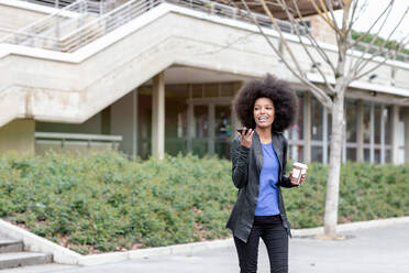 Junge Frau mit Afro-Haar in der Stadt, die spazieren geht und mit ihrem Smartphone spricht - CUF51474