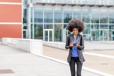 Junge Frau mit Afrohaar in der Stadt, die ein Smartphone mit Touchscreen benutzt - CUF51475