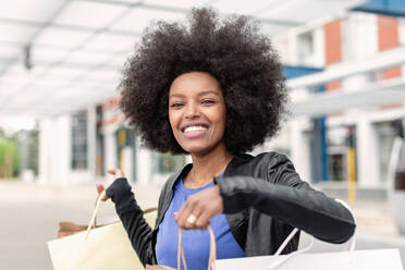 Young woman with afro hair at city train station holding up shopping bags, portrait - CUF51478