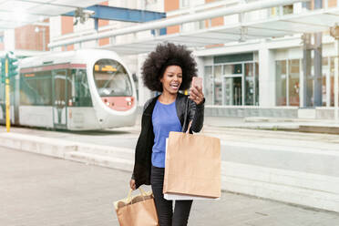 Junge Frau mit Afrofrisur an einer S-Bahn-Station, die Einkaufstüten trägt und ein Smartphone-Selfie macht - CUF51479