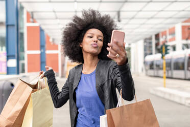 Young woman with afro hair at city train station carrying shopping bags, puckering lips for smartphone selfie - CUF51480