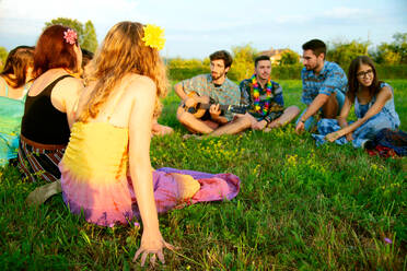 Große Gruppe junger Erwachsener, die auf einem Feld sitzen und einer Akustikgitarre lauschen - CUF51519