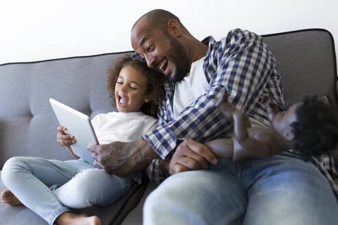 Happy father and daughter sitting on couch at home looking at tablet - JPTF00187