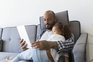 Father and daughter sitting on couch at home together looking at tablet - JPTF00188