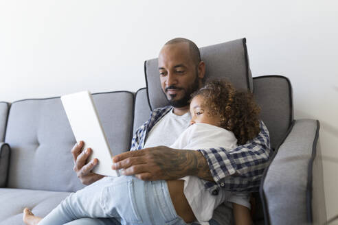 Vater und Tochter sitzen zu Hause auf der Couch und schauen auf ein Tablet - JPTF00188