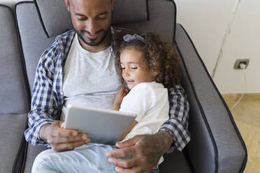 Father and daughter sitting on couch at home together looking at tablet - JPTF00189