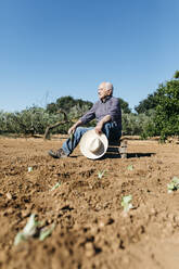 Senior farmer sitting on wooden box, after planting vegetables in the garden - JRFF03397