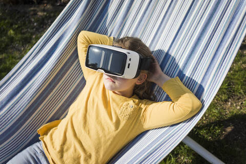 Mädchen liegt in Hängematte im Garten und trägt VR-Brille, lizenzfreies Stockfoto