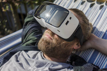 Mann liegt in Hängematte im Garten und trägt VR-Brille - MOEF02277