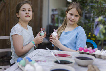 Porträt von zwei Mädchen, die auf einem Gartentisch Ostereier verzieren - MOEF02303
