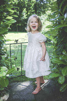 Porträt eines glücklichen kleinen Mädchens im weißen Kleid, das am Gartentor steht - IHF00145