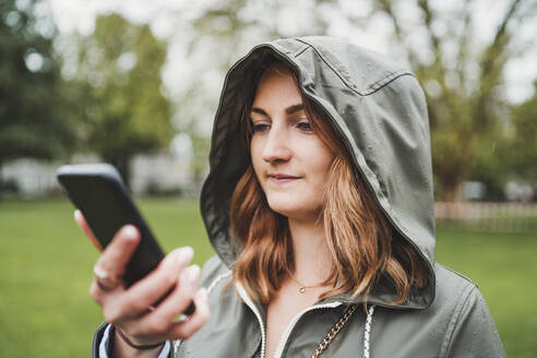 Junge Frau in Kapuzenjacke und mit Handy an einem regnerischen Tag - FBAF00776