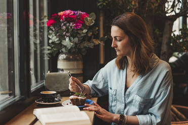 Junge Frau mit einem Buch isst einen Kuchen in einem Café - FBAF00785