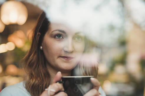 Junge Frau in einem Café trinkt Kaffee hinter einer Fensterscheibe - FBAF00789